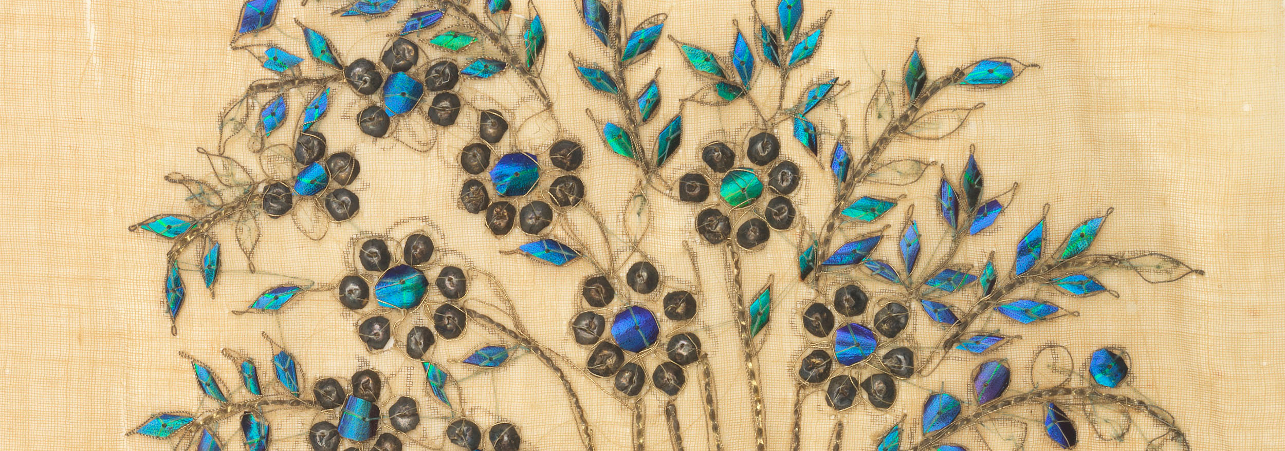A close-up shot of a detail from a textile showing several fragments of iridescent beetle wings sewn onto the fabric.