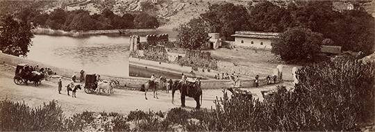 A black and white photograph of the Amber Fort, Jaipur, depicting bullock carts, camels and an elephant near a waterbody.