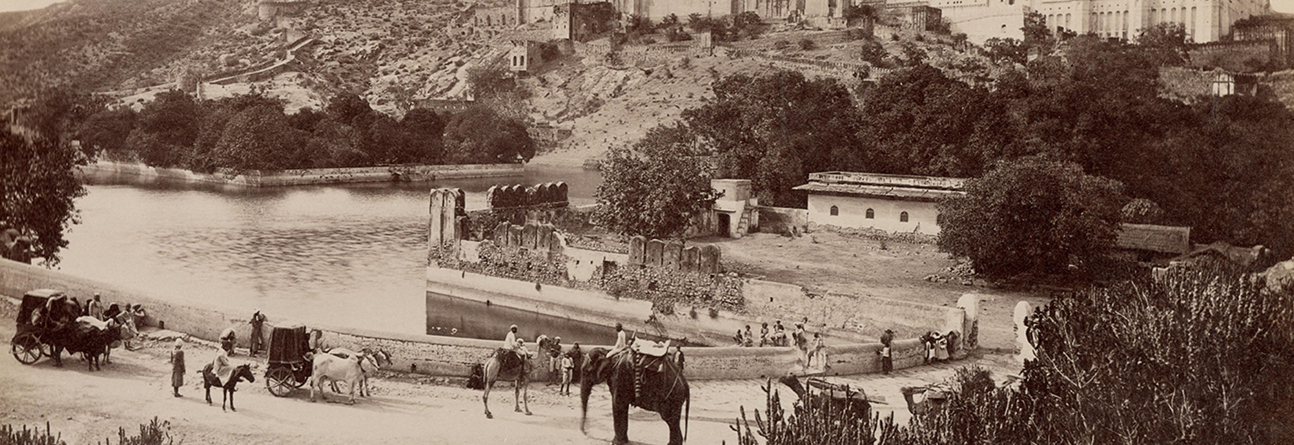 A black and white photograph of the Amber Fort, Jaipur, depicting bullock carts, camels and an elephant near a waterbody.