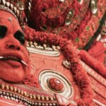 A Theyyam performer dressed in costume, with facial make-up and metal fangs protruding from his mouth.