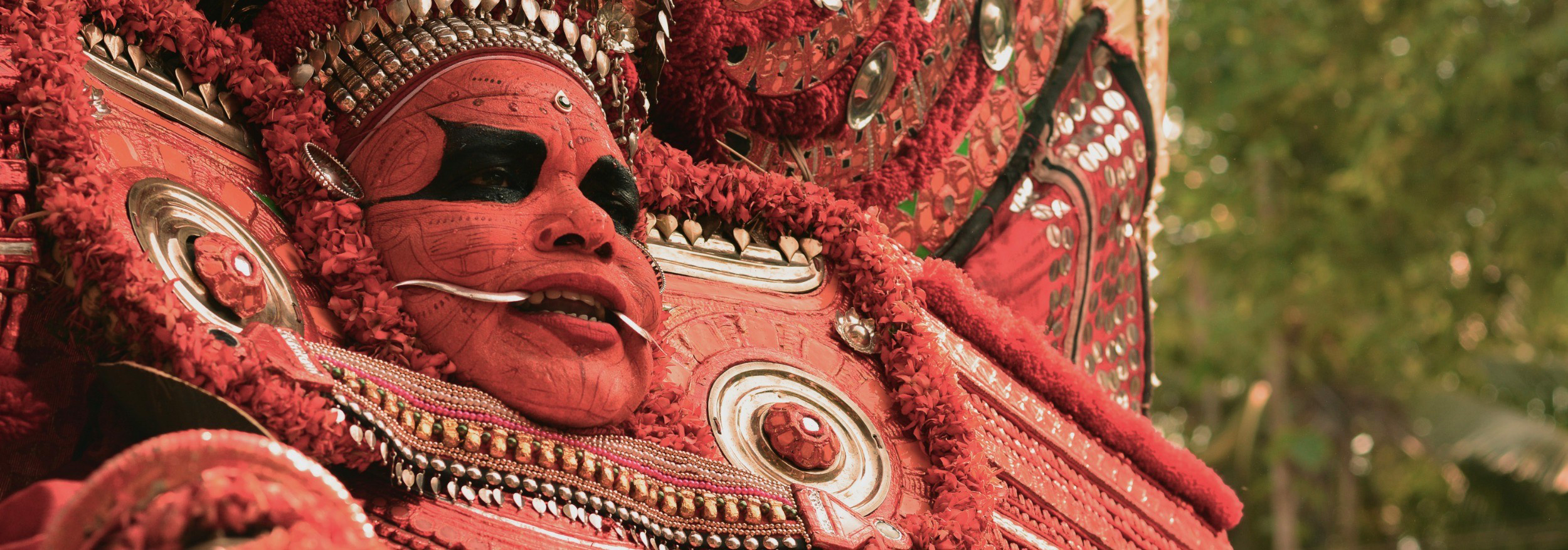 A Theyyam performer dressed in costume, with facial make-up and metal fangs protruding from his mouth.
