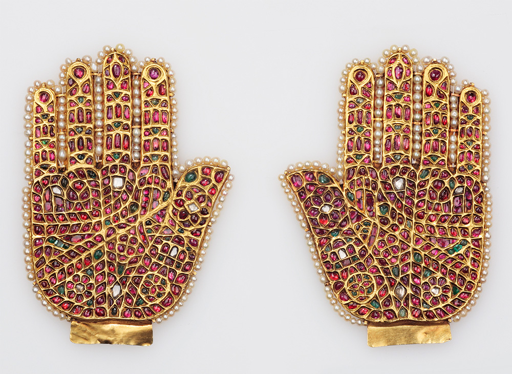 A pair of gold metallic hands infilled with an arrangement of rubies, emeralds and diamonds outlined by a string of pearls.