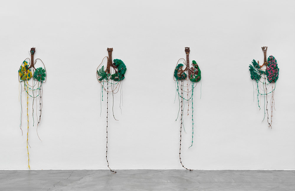 Four lung-shaped objects made of loose wire and thread resembling inverted trees being displayed on a white wall.