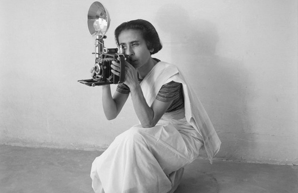A woman dressed in a white sari holding up a camera at eye level while kneeling on the ground