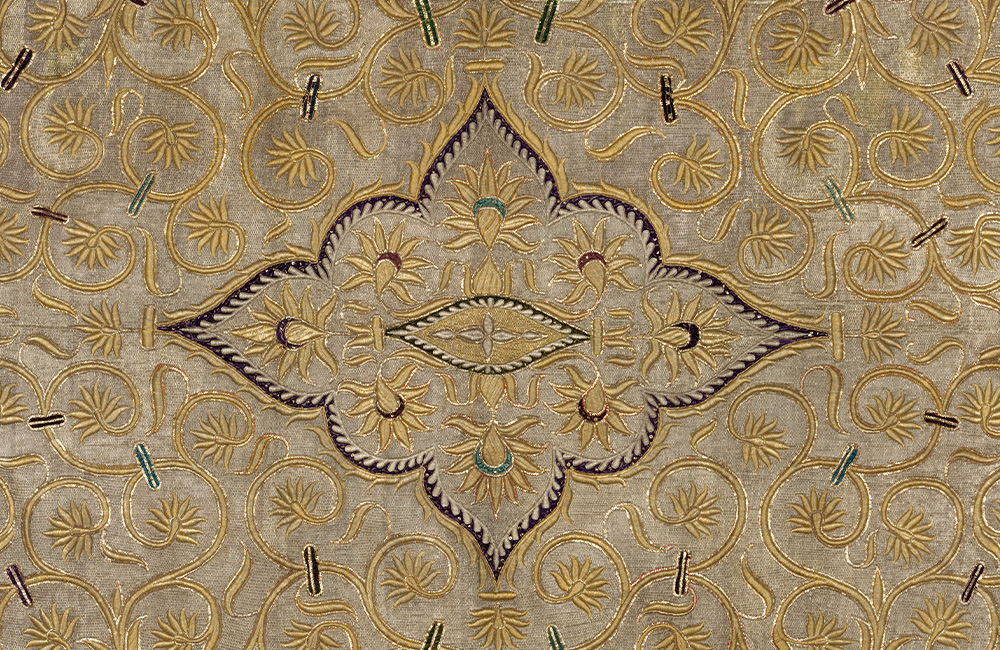 A beige textile featuring embroidered floral scrolls and a central cartouche infilled with petalled flowers.