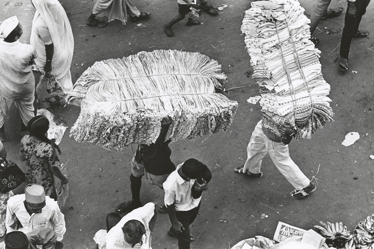 A scene from a market with hawkers carrying large stacks of bundled cloth over their heads.