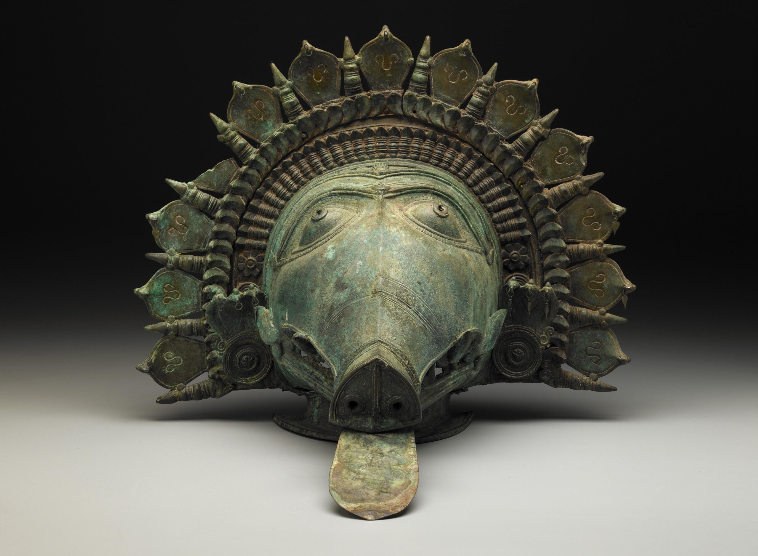 A metal Bhuta mask with the image of a boar, with a protruding tongue, encircled by a partial halo.