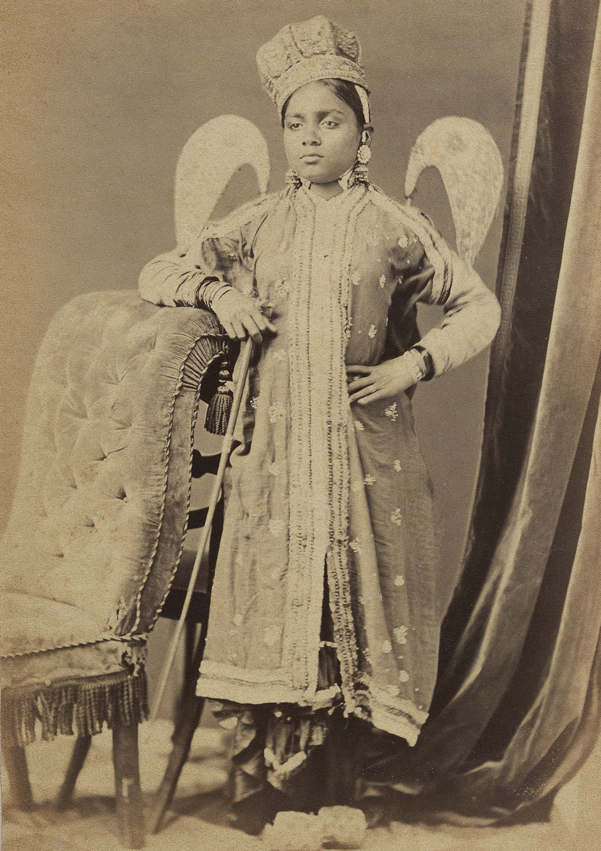 A sepia-toned photograph of a woman dressed as a winged fairy, standing upright next to a chair.