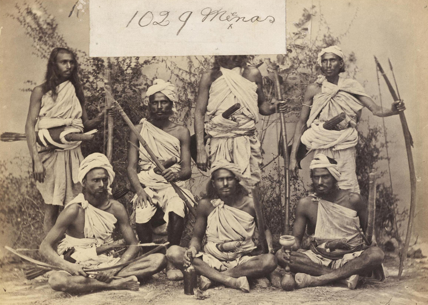 A sepia-toned group photograph of men from the Meena tribe dressed in white cloth and bearing bows and arrows.