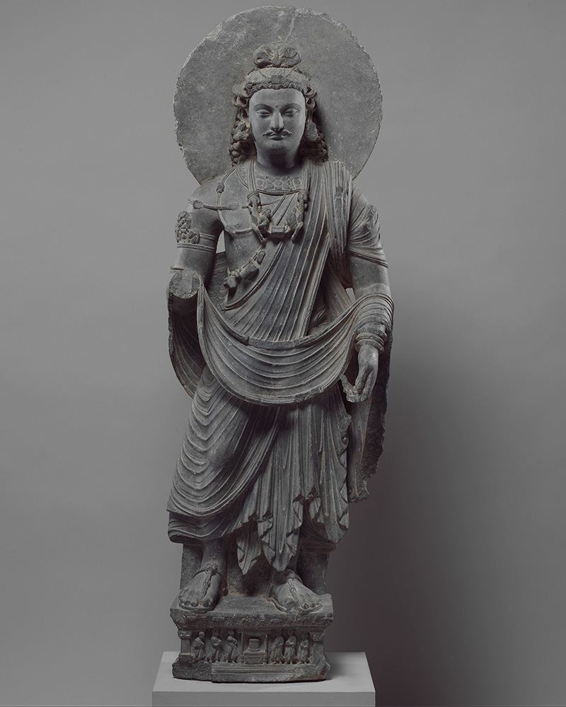 A schist statue of the Bodhisattva Maitreya draped in cloth, wearing jewellery and surrounded by a halo.