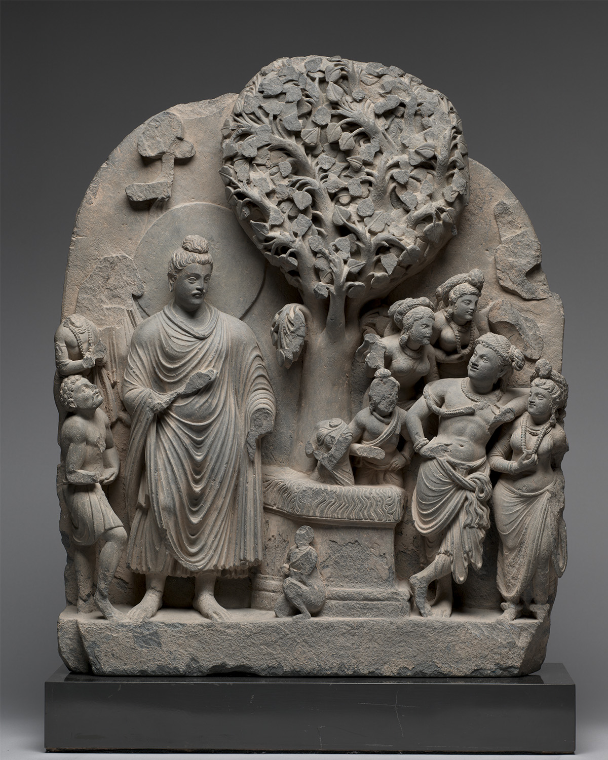 A stone relief of the Bodhi Tree with Siddhartha on the left and flanked by villagers, and the earth goddess at the bottom.