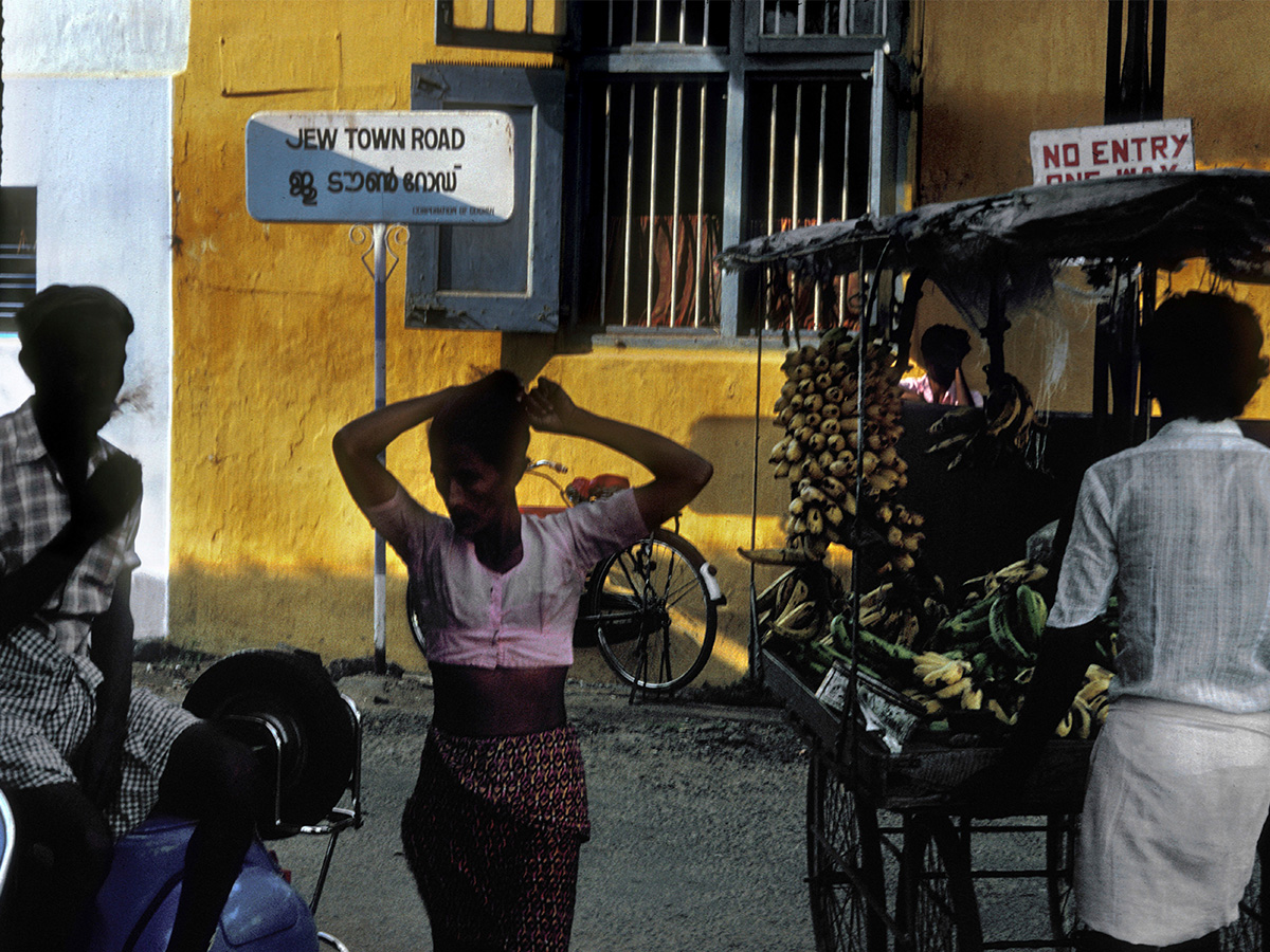 A photograph of the Jew Town Road in Kochi, with a fruit seller and passersby.