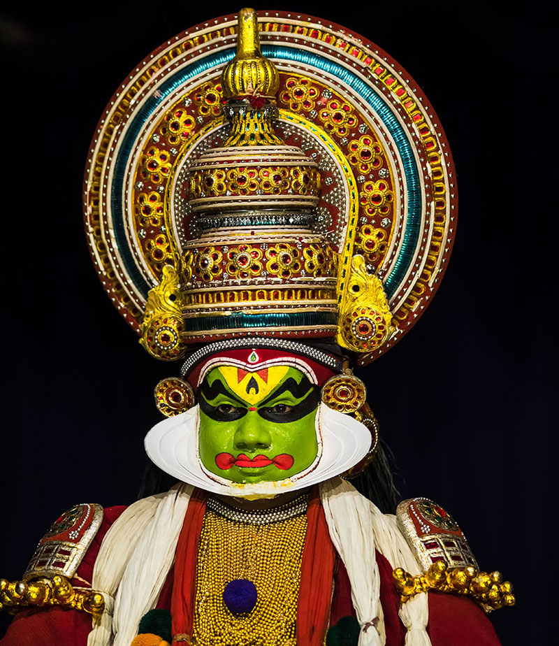 A Kathakali dancer in the costume of a male character, with a tall headdress, green facial make-up and a white facial mask.