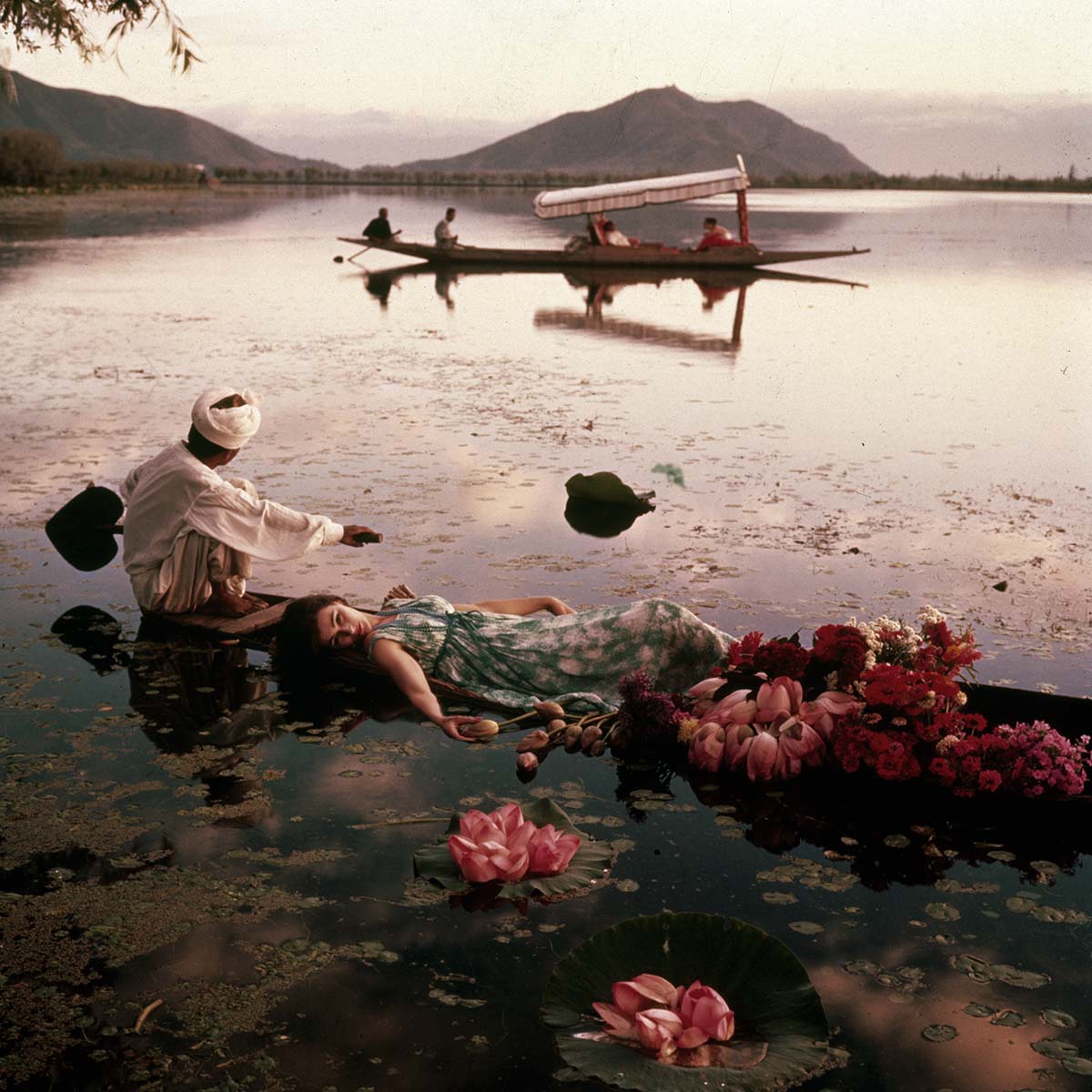 A photograph of a model lying on a boat full of flowers, ferried by a rowman in the Dal Lake, blooming with lotuses.