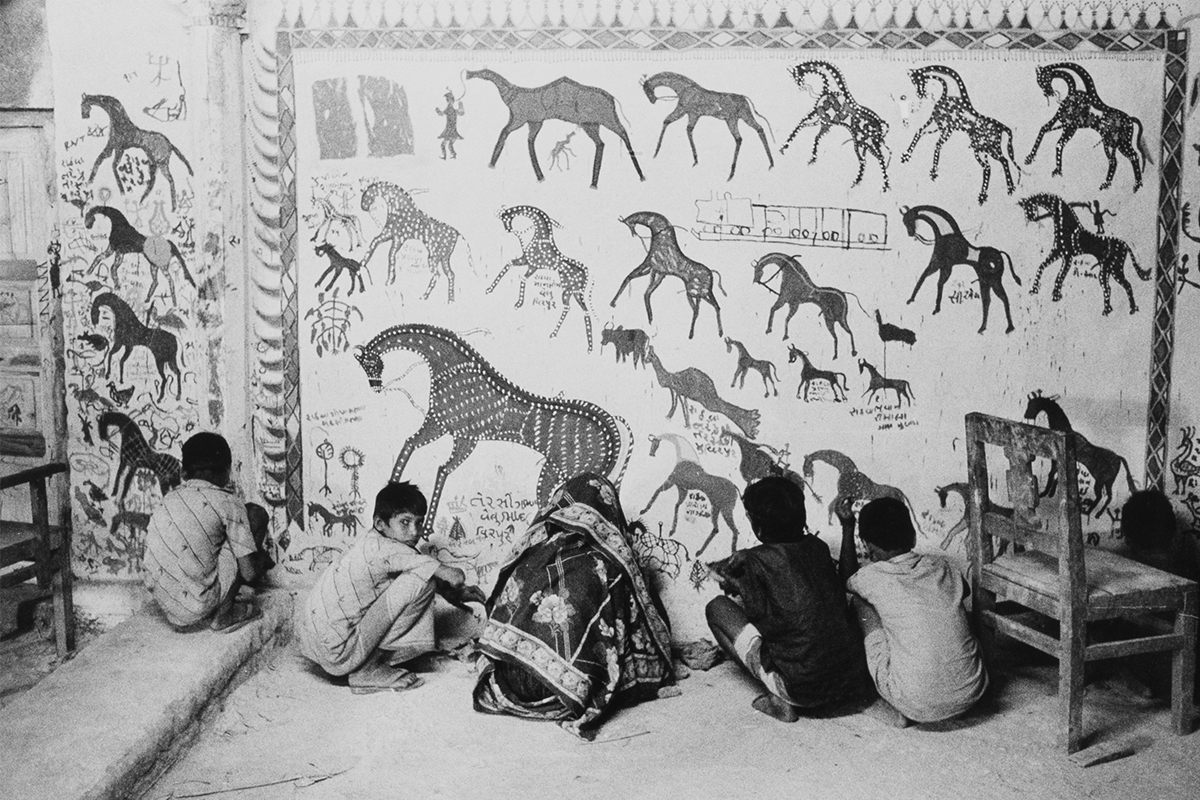 A photograph of a woman and several children looking at a Pithora mural, with one of the children facing the camera.