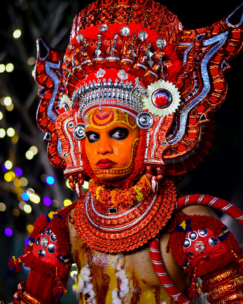 A Theyyam performer wearing a large red head-dress, a red costume and orange facial make-up.