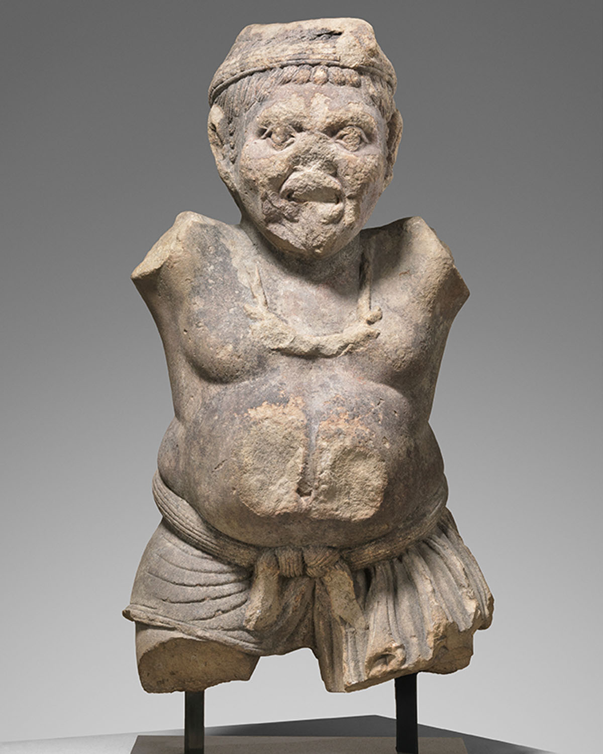 A stone sculpture with disfigured facial features, a protruding belly and missing limbs.