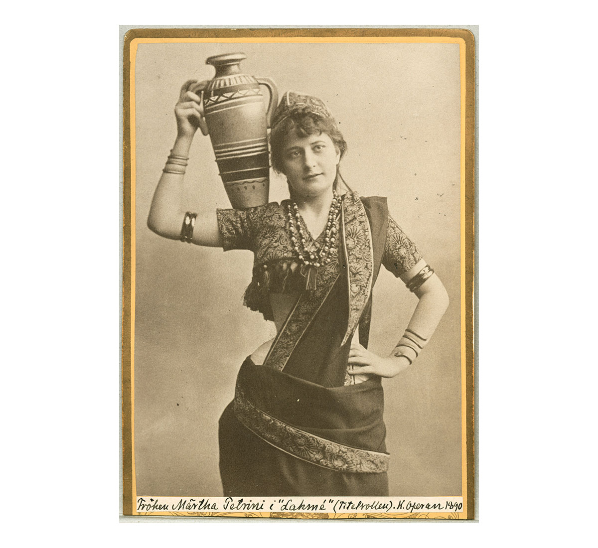 A photograph of a woman in a saree and blouse holding up an urn on her right shoulder.