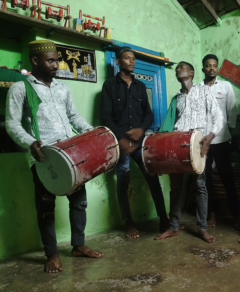 Photograph of four men standing, two men playing drums, inside the Bava Gor shrine in Gujarat