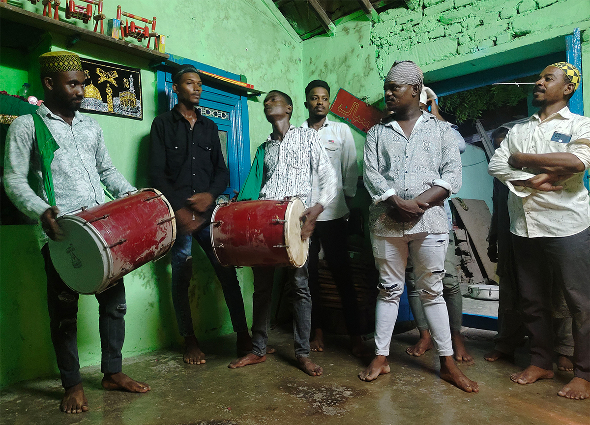 Photograph of eight men standing, two men playing drums, inside the Bava Gor shrine in Gujarat